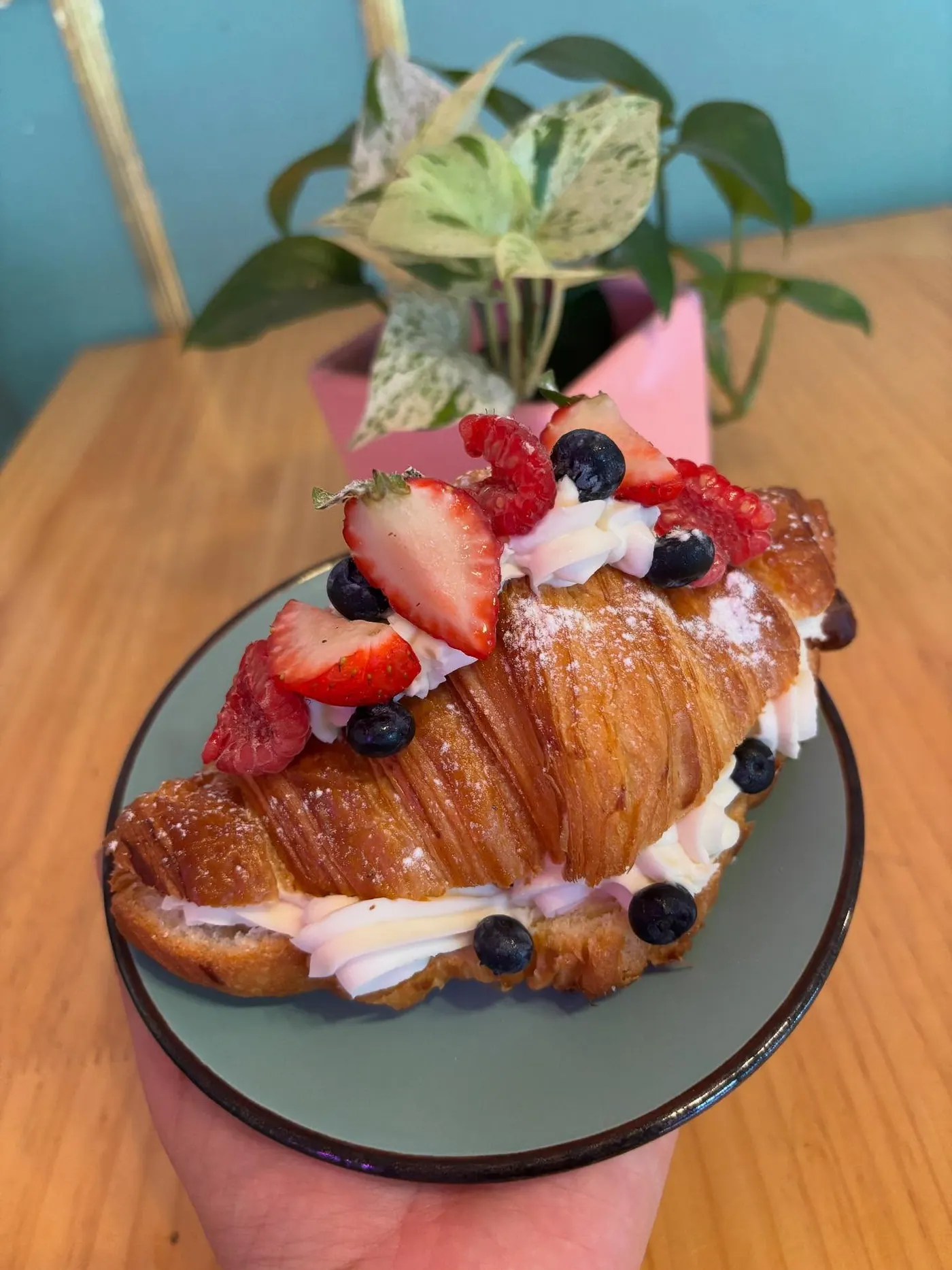 Croissant relleno de crema con fresas y arándanos, sobre la pared rosa del local